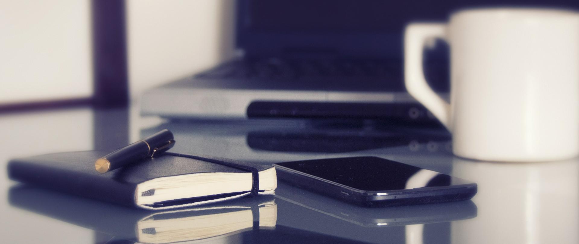 notes i długopis, w tle telefon, komputer i kubek do picia