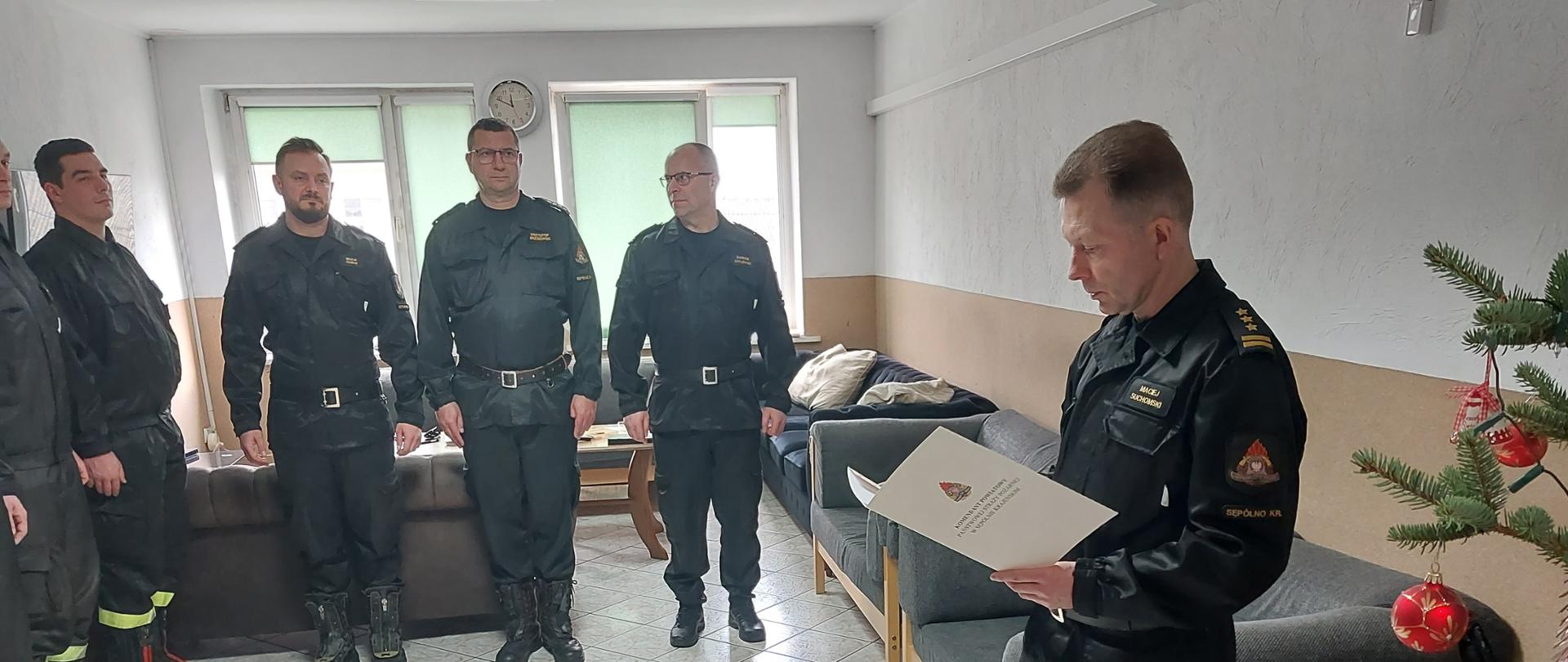 Zdjęcie wykonano w budynku Komendy Powiatowej Państwowej Straży Pożarnej w Sępólnie Krajeńskim w pomieszczeniu świetlicy. Strażacy Państwowej Straży Pożarnej w ubraniach koszarowych stoją w szeregu, a Komendant Państwowej Straży Pożarnej starszy brygadier Maciej Suchomski odczytuje decyzję o awansie jednego ze strażaków.