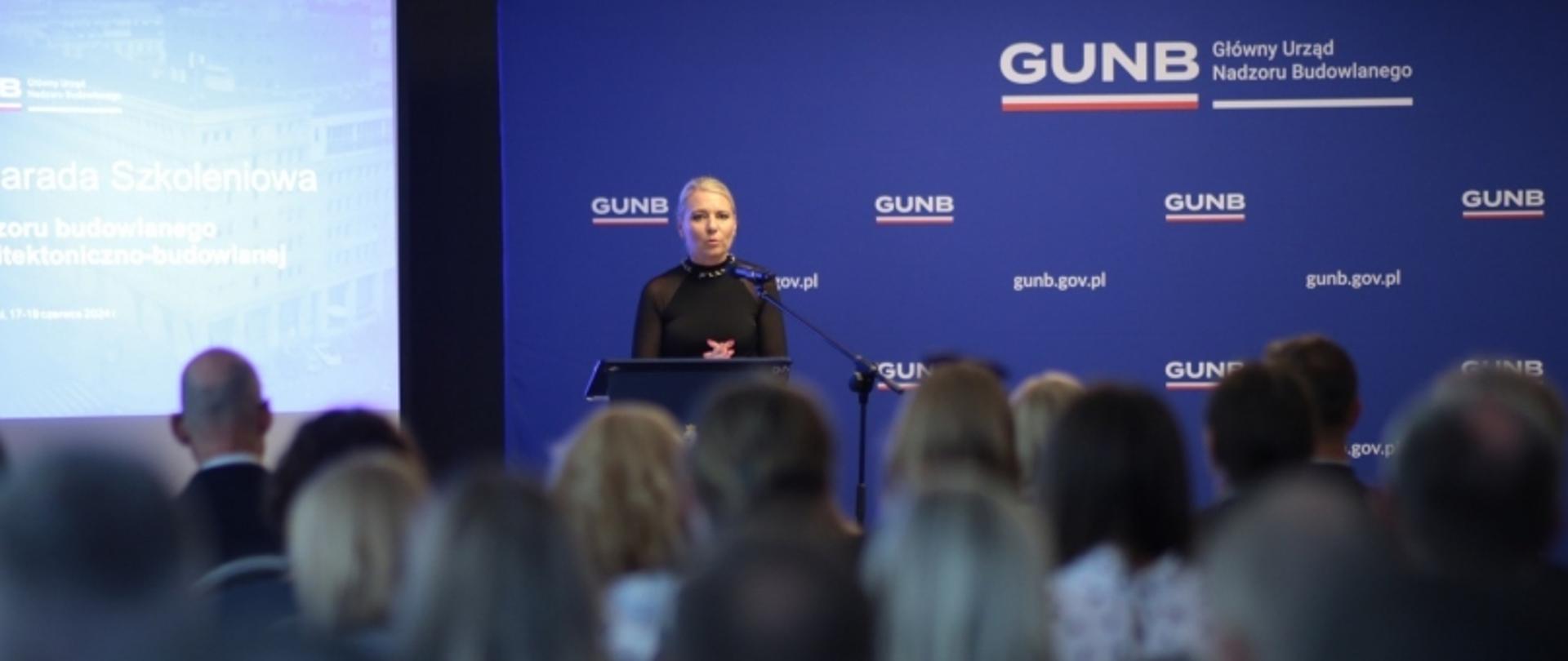 Osoba przemawiająca przy mównicy podczas narady szkoleniowej GUNB, z ekranem i banerem „gunb.gov.pl” w tle.
