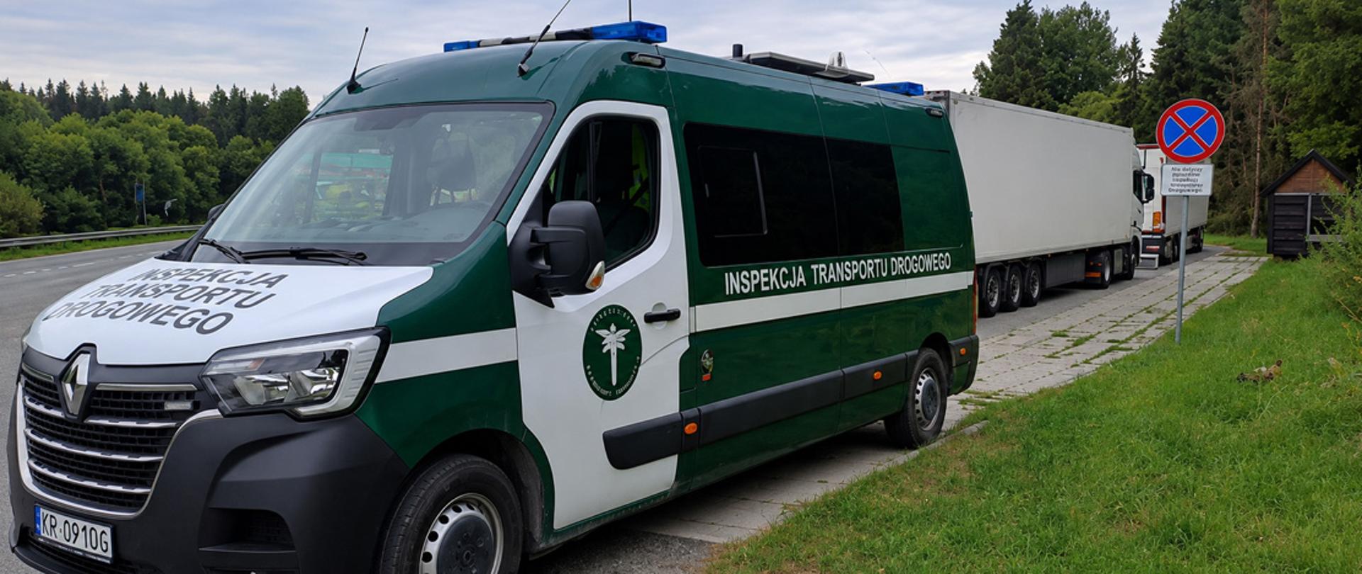 Zielono-biały samochód ITD z napisem Inspekcja Transportu Drogowego stoi przy drodze obok kontrolowanej ciężarówki. W tle zalesiony teren.