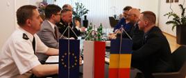 Na przedzie u dołu widoczne trzy flagi. Unii Europejskiej, Polska oraz Rumuńska stojące na stole. W głębi czterech mężczyzn po lewej stronie stołu oraz kobieta i dwaj mężczyźni siedzący naprzeciw nich. 