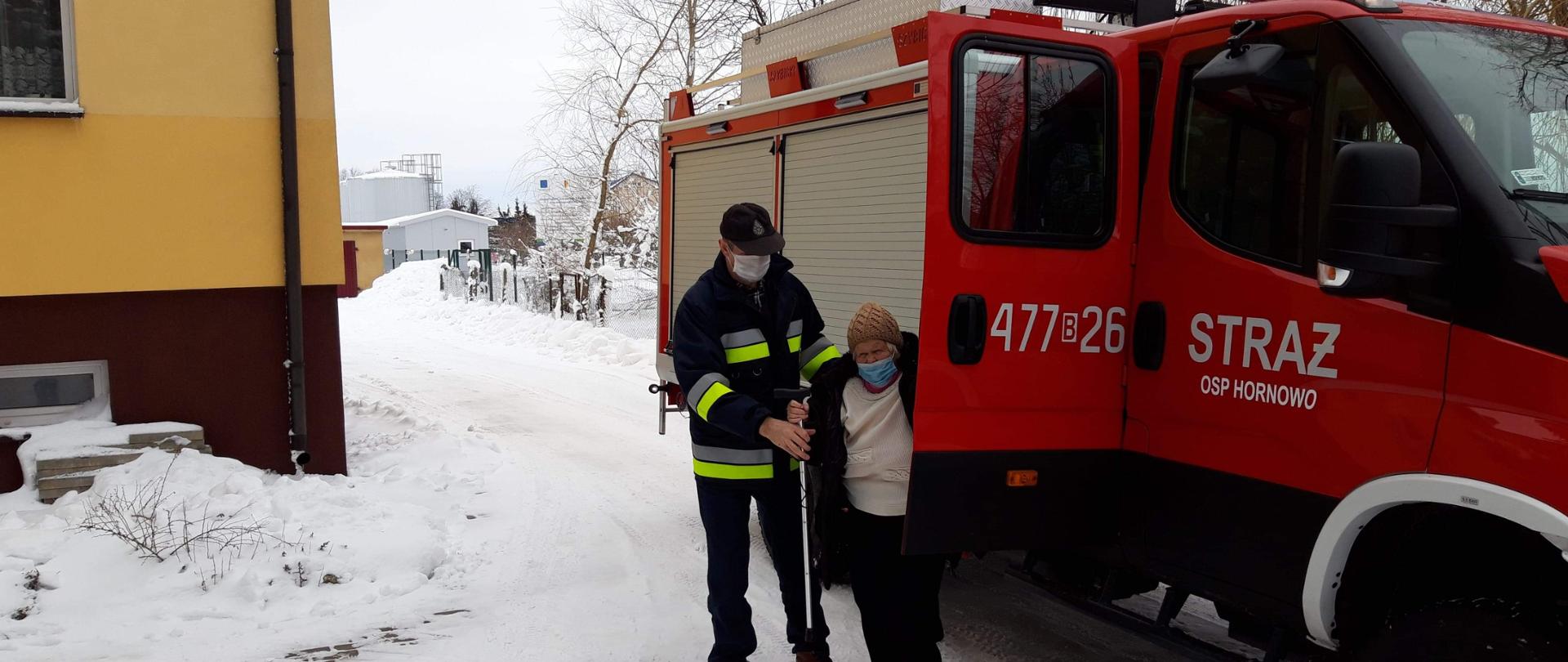 Czerwony samochód pożarniczy, zaparkowany na śniegi. Obok stoi strażak oraz osoba starsza która jest zabierana na sczepienie.