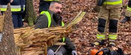 Zdjęcie przedstawia zajęcie z cięcia pilarką mechaniczną w lesie na zdjęciu strażak tnący oraz instruktor nadzorujący.