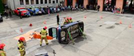 Na środku placu leży przewrócony na bok granatowy samochód osobowy, obok strażacy ubrani w ubrania specjalne prowadza pokazowa akcje ratownicza. Z tyłu przed garażami i budynkiem zgromadzeni ludzie obserwują pokaz ratownictwa.