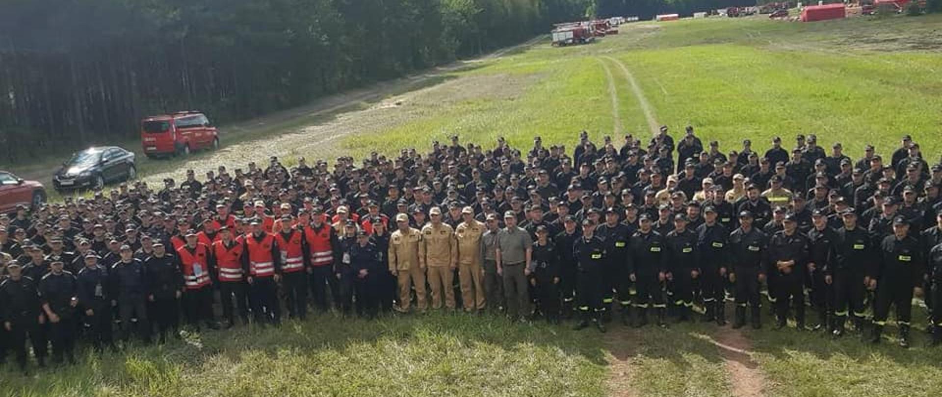 Na zdjęciu widzimy grupę ok. 400 strażaków pozującą do zdjęcia w scenerii lasu. W tle widać pojazdy pożarnicze oraz obozowisko.