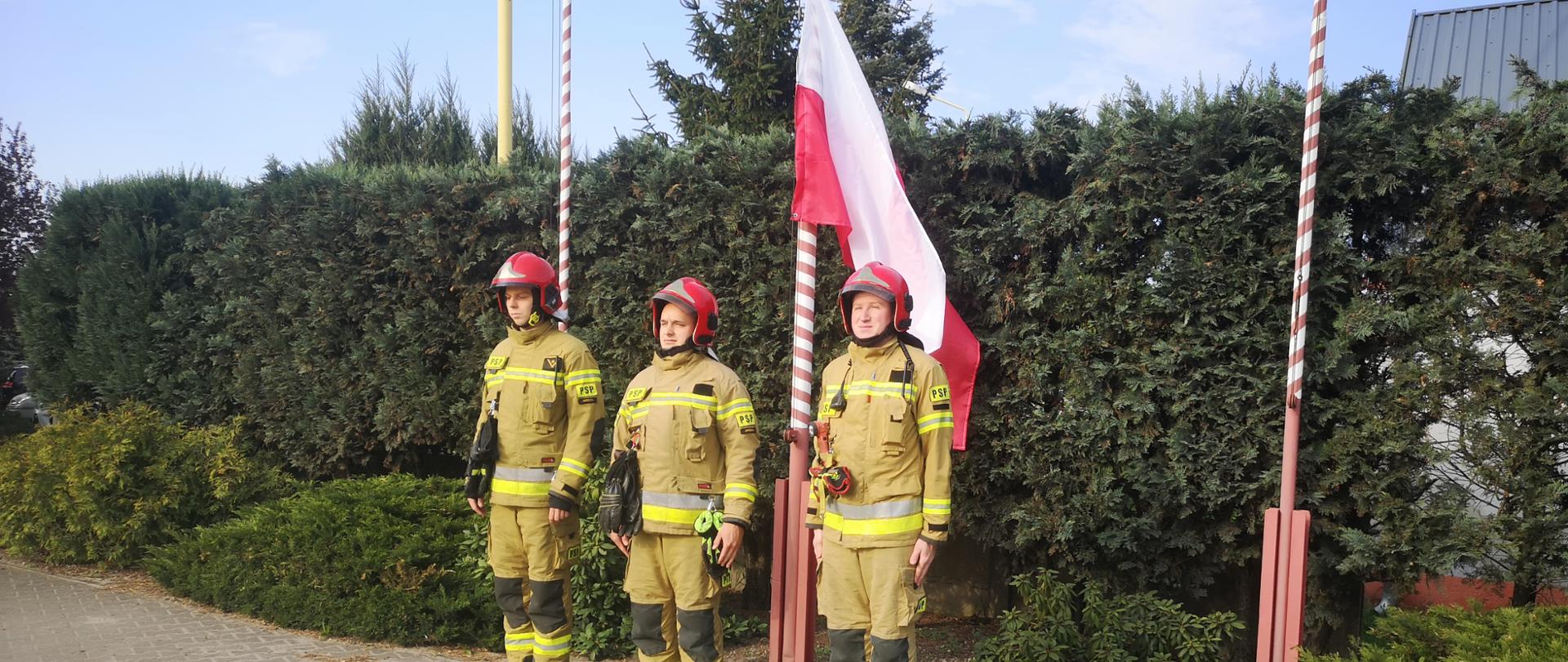 Trzech funkcjonariuszy w umundurowaniu specjalnym stoją pod masztem flagowym.