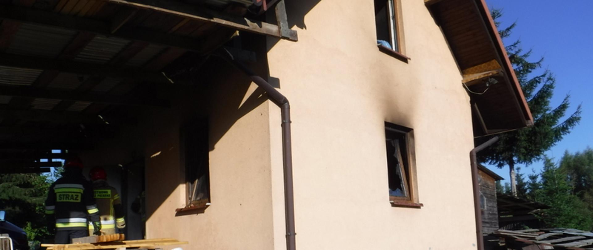 Zdjęcie przedstawia murowany budynek gospodarczy w którym miał miejsce pożar. Ściany budynku pomalowane są beżową farbą z widocznymi okopceniami nad oknem. Przed budynkiem znajduje się drewno opałowe, garnki i wiadra. Do budynku wchodzą dwaj strażacy.