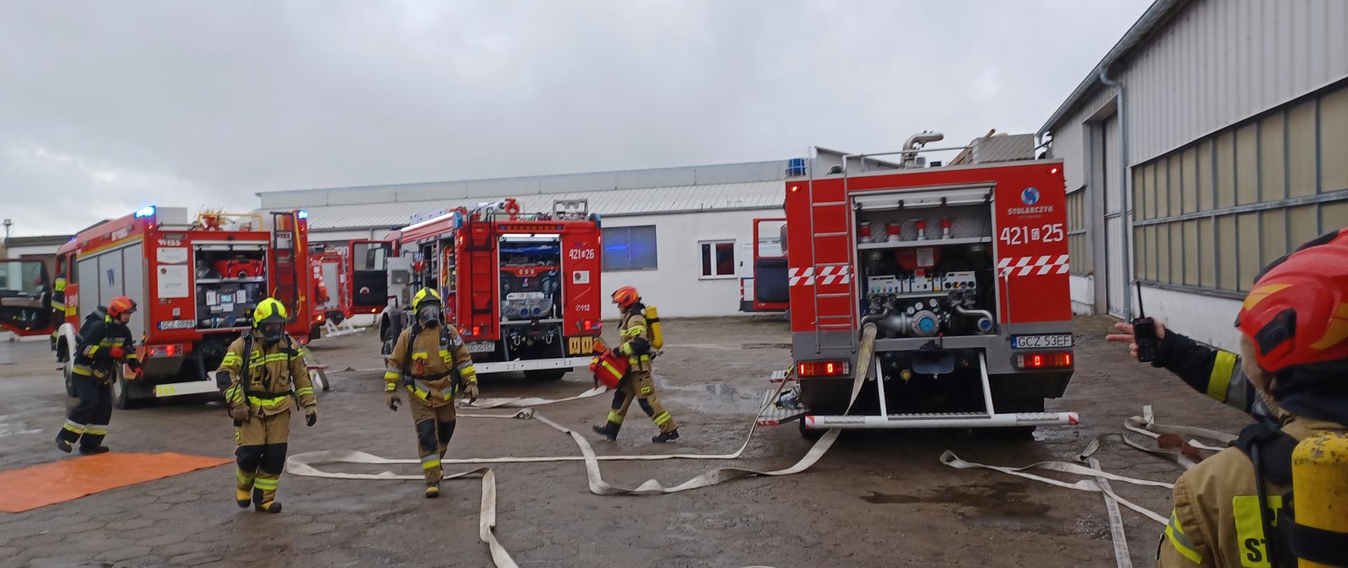 Na pierwszym planie widać strażaków w sprzęcie ochrony układu oddechowego, rozwinięte węże pożarnicze i strażaków sprawiających sprzęt. W tle stoją samochody pożarnicze. 