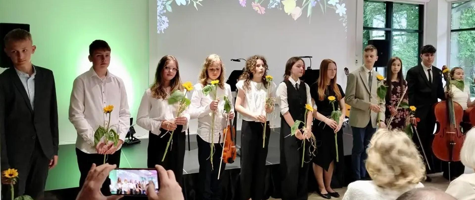 Grupa młodzieży stoi w rzędzie na scenie po koncercie, część trzyma kwiaty i instrumenty smyczkowe. W tle ekran z kwiatowym motywem, a publiczność z pierwszego rzędu robi im zdjęcia telefonem.