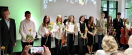 Grupa młodzieży stoi w rzędzie na scenie po koncercie, część trzyma kwiaty i instrumenty smyczkowe. W tle ekran z kwiatowym motywem, a publiczność z pierwszego rzędu robi im zdjęcia telefonem.