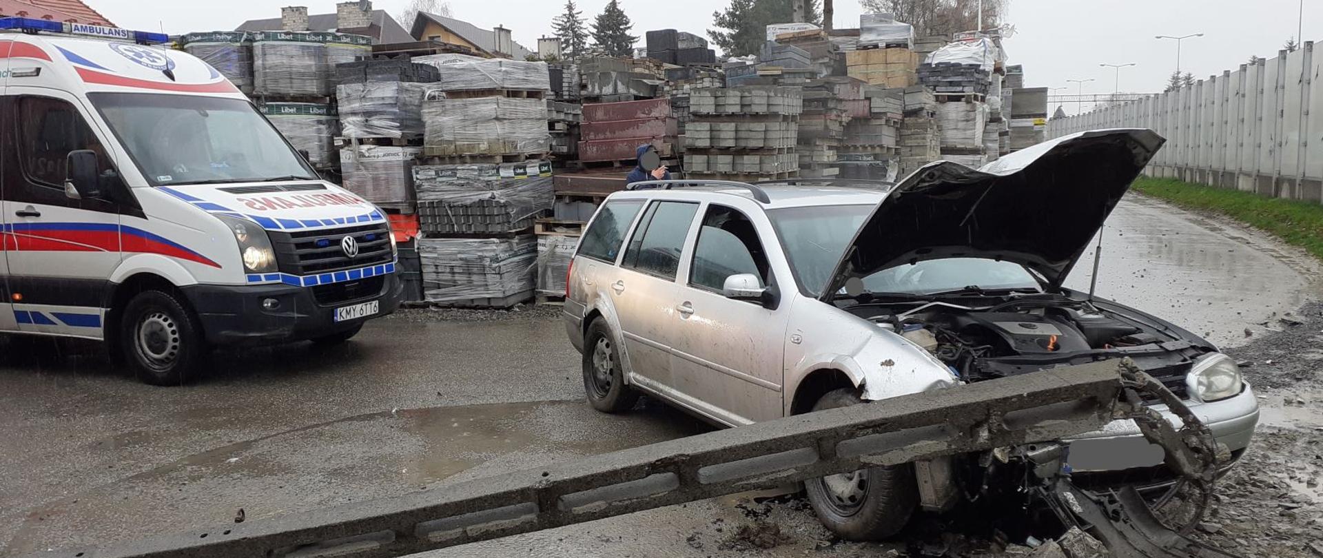 Na zdjęciu widoczny samochód osobowy w kolorze srebrnym, który uderzył w betonowy słup energetyczny. W aucie otwarta jest pokrywa silnika. Zniszczony słup leży przed samochodem w poprzek jezdni. Z lewej strony częściowo widoczna karetka pogotowia ratunkowego. W tle zdjęcia skład materiałów budowlanych (kostka brukowa).