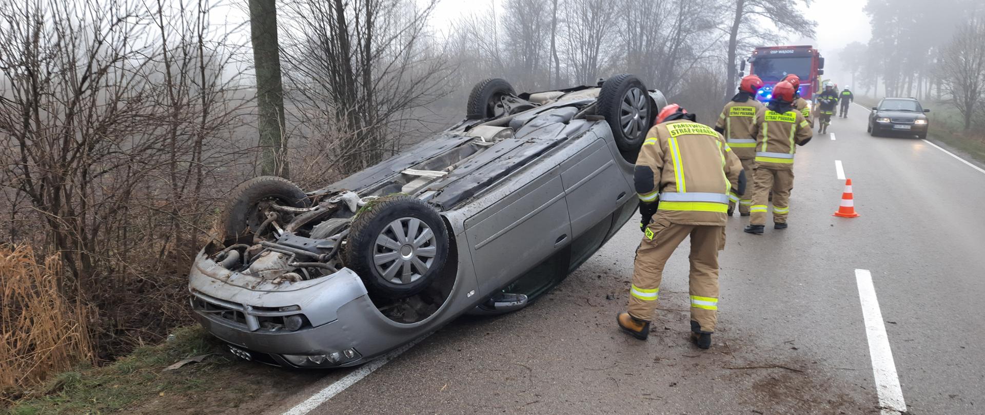 Na zdjęciu samochód osobowy przewrócony na dach leży na jezdni przy nim strażak PSP w ubraniu specjalnym zagląda do wnętrza pojazdu. W drugim planie trzech strażaków PSP w ubraniach specjalnych odwróconych tyłem. W tle samochód strażacki GBA oraz samochod osobowy cywilny, pomiędzy nimi strażacy OSP w ubraniach specjalnych.