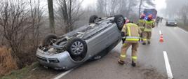 Na zdjęciu samochód osobowy przewrócony na dach leży na jezdni przy nim strażak PSP w ubraniu specjalnym zagląda do wnętrza pojazdu. W drugim planie trzech strażaków PSP w ubraniach specjalnych odwróconych tyłem. W tle samochód strażacki GBA oraz samochod osobowy cywilny, pomiędzy nimi strażacy OSP w ubraniach specjalnych.