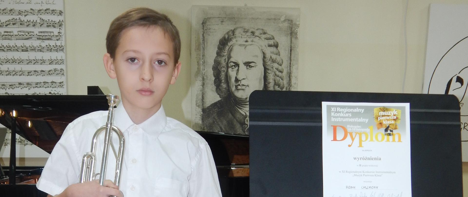  Na zdjęciu stoi chłopiec trzyma trąbkę obok ustawiono pulpit z Dyplomem z napisem Wyróżnienie. W tle fortepian i portret Jana Sebastiana Bacha.