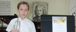 Na zdjęciu stoi chłopiec trzyma trąbkę obok ustawiono pulpit z Dyplomem z napisem Wyróżnienie. W tle fortepian i portret Jana Sebastiana Bacha.