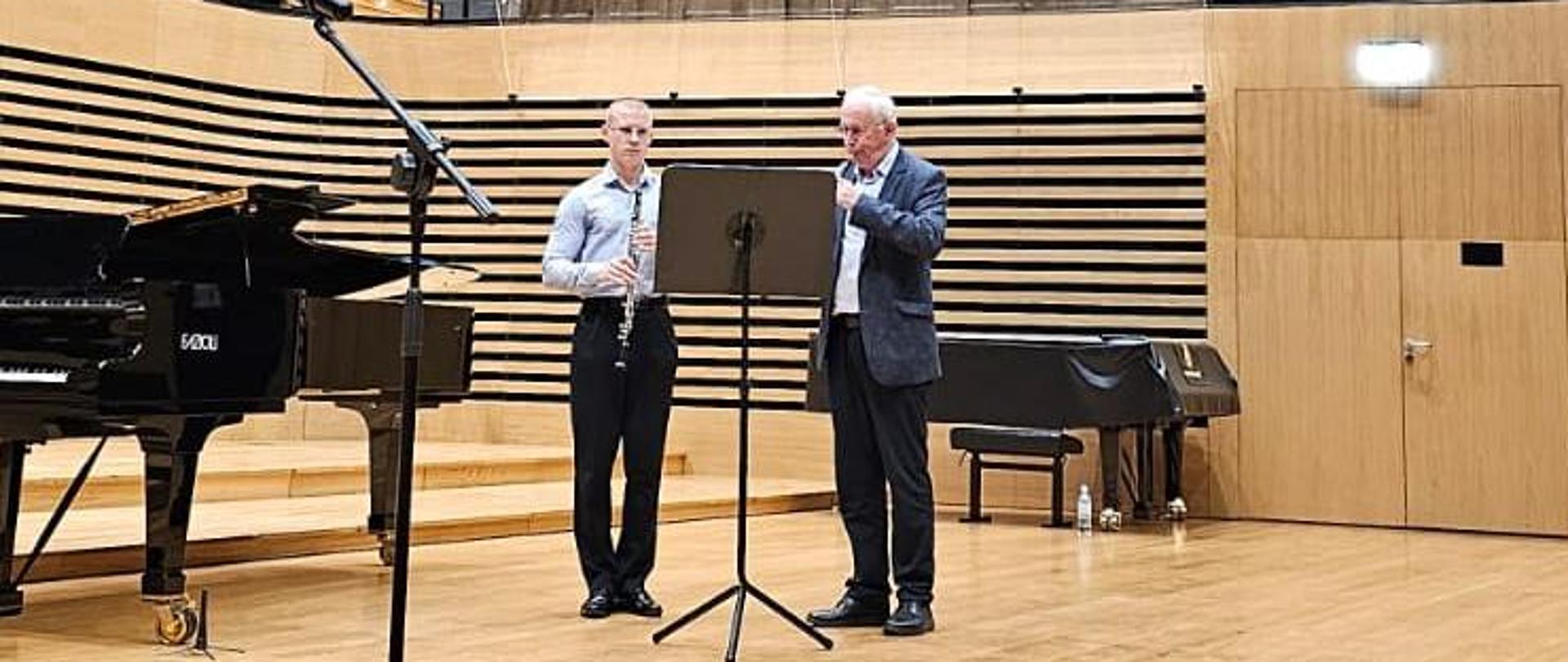 Mężczyzna i nastolatek stoją obok siebie przy pulpicie na estradzie sali koncertowej. Mężczyzna gra na oboju, chłoak trzyma obój przed sobą.