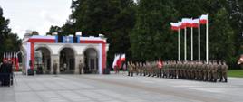 Zdjęcie przedstawia Grób Nieznanego Żołnierza na pl. Marszałka Józefa Piłsudskiego w Warszawie. Po prawej stronie zdjęcia stoją ustawieni w szpaler żołnierze Wojska Polskiego, zaś tuż za nimi 5 biało-czerwonych flag, powiewających na wysokich masztach. W tle ciemnozielone kępy drzew. 