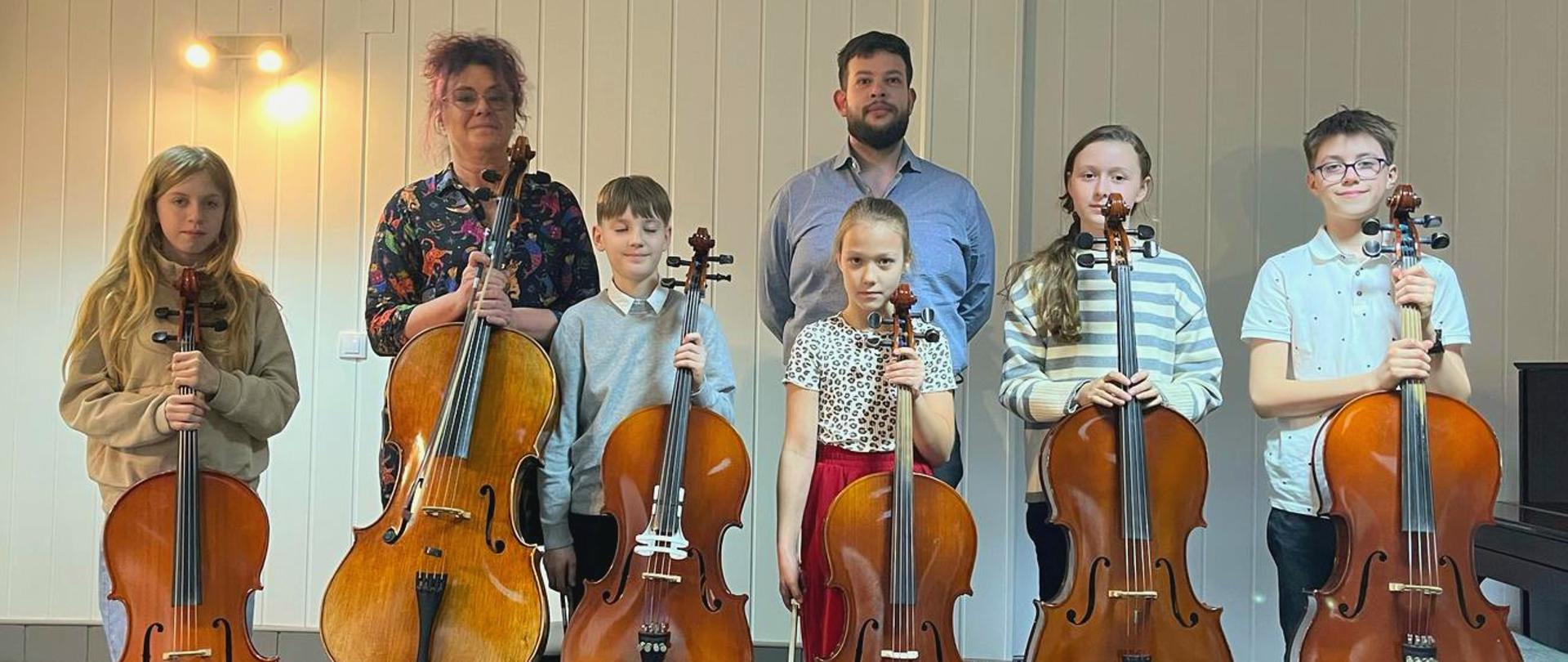 kilkoro stojących dzieci trzymających wiolonczele, za nimi dwóch pedagogów, obok po prawej pianino, białe tło