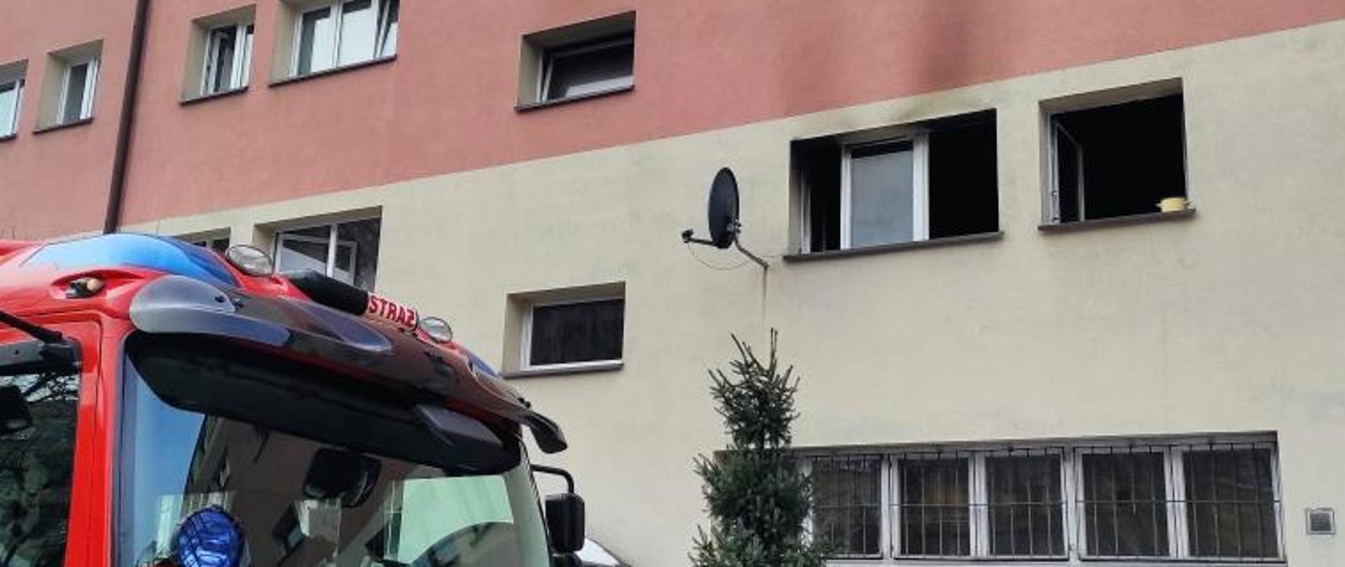 na pierwszym planie widać przód pojazdu gaśniczego, na drugim planie widać okopconą elewację bloku nad oknami jednego z mieszkań
