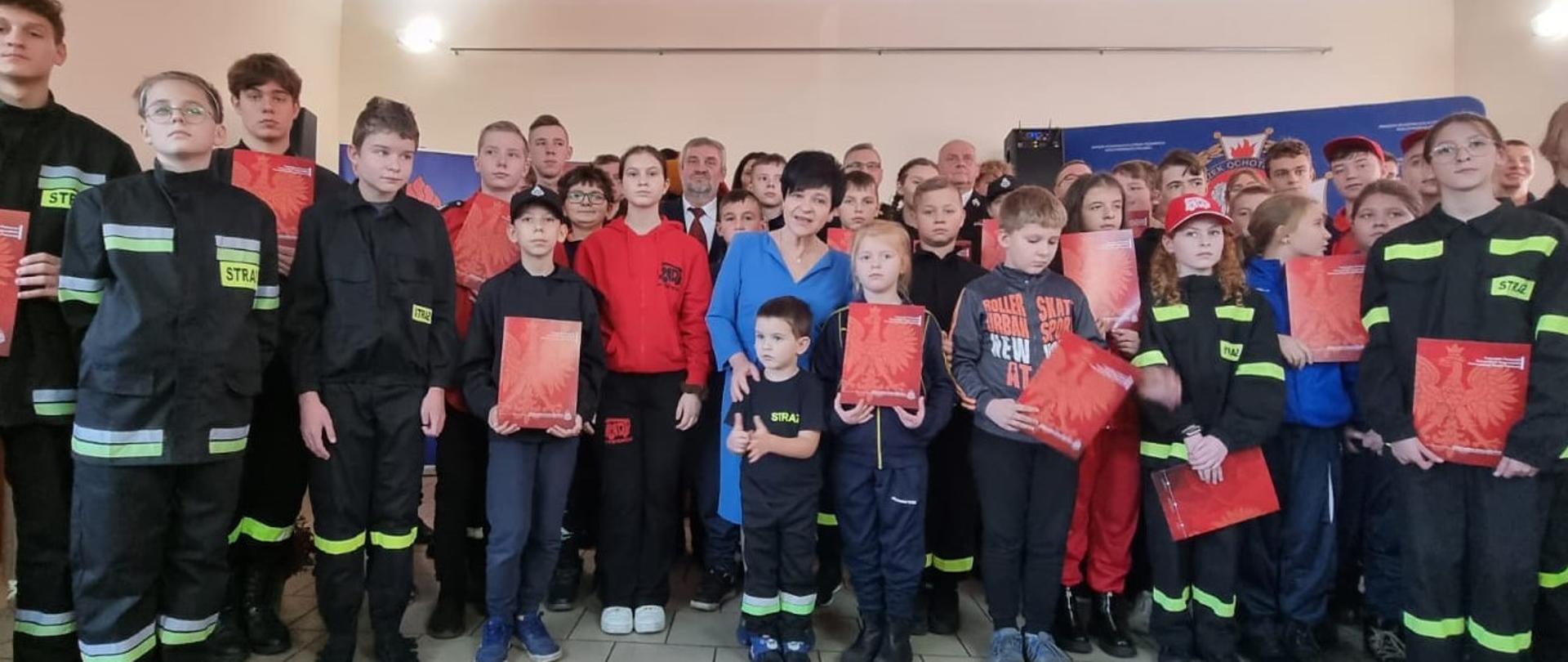 Zdjęcie grupowe uczestników wydarzenia, na którym szczególnie widać młodzież strażacką. Są w mundurkach, niektórzy w rekach trzymają otrzymane promesy. Towarzyszą im parlamentarzyści.