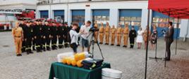 Grupa strażaków ustawionych w szeregu podczas uroczystej zbiórki na placu. Na środku operator kamery oraz fotograf.