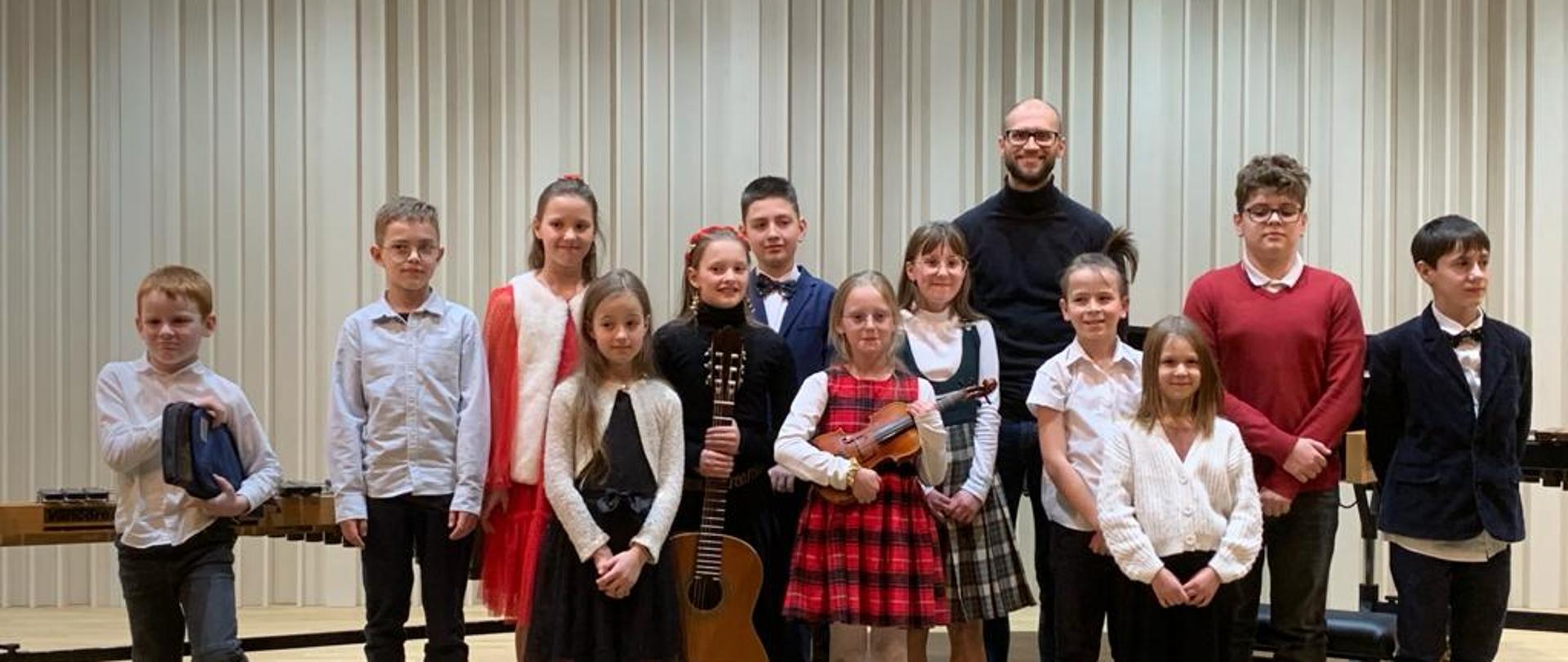 Na zdjęciu grupa uczniów, którzy wystąpili podczas koncertu klasy perkusji. Z tyłu za uczniami mężczyzna w czarnym golfie, lekkim zarostem i okularach.