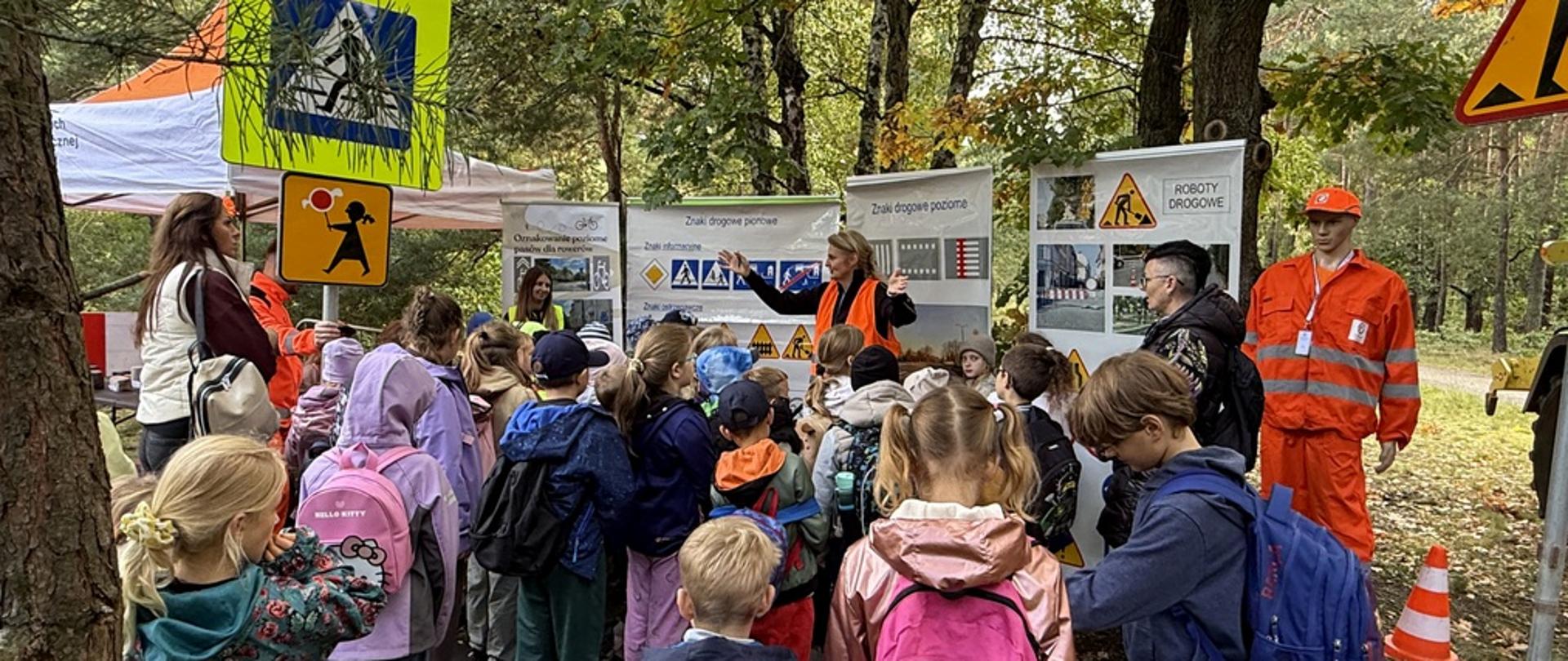 Dzieci zgromadziły się wokół instruktorki w pomarańczowej kamizelce, która tłumaczy zasady ruchu drogowego. W tle stoją tablice informacyjne ze znakami drogowymi oraz napisami, m.in. „Znak drogowy pionowy” i „ROBOTY DROGOWE”. Nad głowami dzieci zawieszono żółty znak drogowy przedstawiający przejście dla pieszych. 