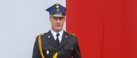 Zdjęcie przedstawia strażaka na tle flagi Polski ubranego w mundur galowy, trzyma w ręku dokumenty.