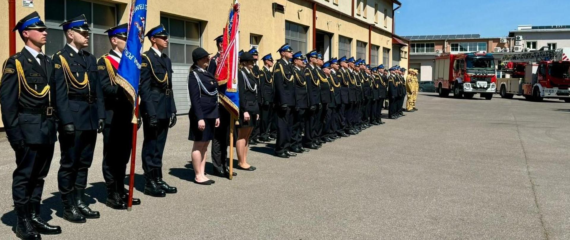 Fotografia wykonana podczas dnia strażaka w komendzie powiatowej Państwowej Straży Pożarnej w Mielcu. Na zdjęciu stoją funkcjonariusze komendy powiatowej PSP w Mielcu na placu przed jednostką ratowniczo-gaśniczą nr 2. Wszyscy strażacy stoją w postawie zasadniczej i są umundurowani w mundury służbowe i galowe. Na pierwszym planie stoi poczet sztandarowy wraz z sztandarem Komendy Powiatowej PSP w Mielcu.