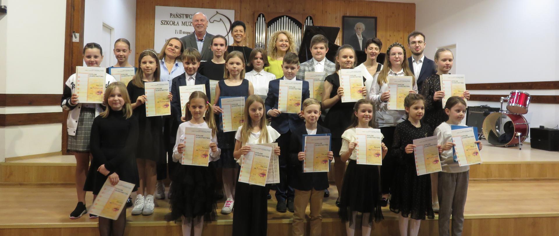 Laureaci konkursu stoją z dyplomami w ręku na scenie PSM w Koninie, w dwóch rzędach, w trzecim rzędzie nauczyciele.