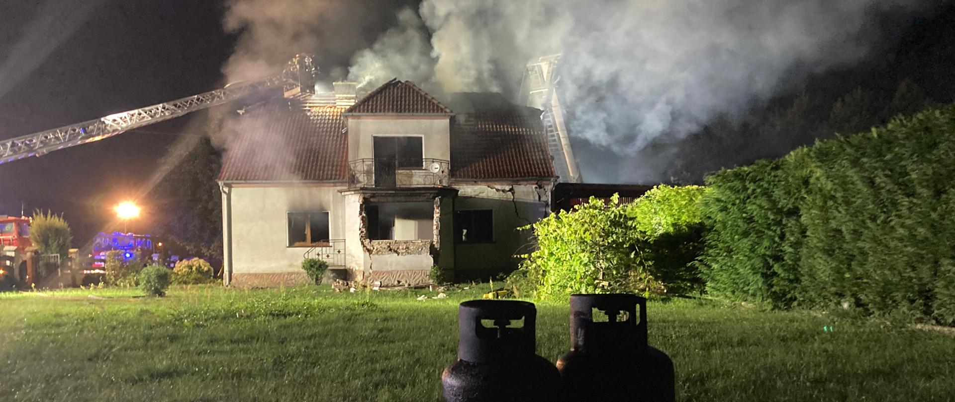 Zdjęcie przedstawia działania strażaków po wybuchu i pożarze w budynku mieszkalnym. Na pierwszym planie wyniesione z obiektu dwie butle gazowe.