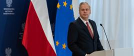 Wiceminister Karol Rabenda stoi za mównicą. W tle ścianka z logotypami MAP i flagi. 
