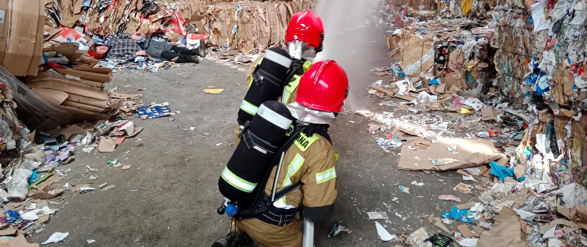 Widok na pomieszczenie hali. Wokół złożona makulatura. Pośrodku dwóch klęczących strażaków ćwiczących gaszenie pożaru.