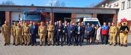 Zdjęcie przedstawia grupę osób pozujących do wspólnej fotografii przed budynkiem remizy strażackiej w słoneczny dzień. W centralnej części stoją mężczyźni w garniturach oraz strażacy w piaskowych mundurach służbowych, a po prawej stronie towarzyszy im maskotka psa w stroju strażaka z dużym czerwonym hełmem. Uczestnicy spotkania są w rzędach na wybrukowanym placu przed garażami jednostki.
W tle, za zgromadzonymi osobami, parkują dwa pojazdy ratownicze: duży, czerwony samochód gaśniczy marki Volvo oraz biały furgon z niebieskimi sygnałami świetlnymi i oznaczeniami straży pożarnej. Budynek posiada liczne bramy garażowe i elewację z elementami czerwonej cegły. Całość kompozycji ma charakter oficjalny i dokumentuje wydarzenie z udziałem przedstawicieli służb mundurowych oraz cywilnych gości.
