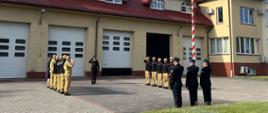 Na zdjęciu widać 15-stu strażaków. 11 z nich stoi w dwóch pięcioosobowych szeregach, zwróconych twarzami do siebie. Między nimi znajduje się strażak (dowódca) zwrócony twarzą do masztu z flagą znajdującym się na przeciw niego. Flagę polski na maszt wciąga 3 strażaków.