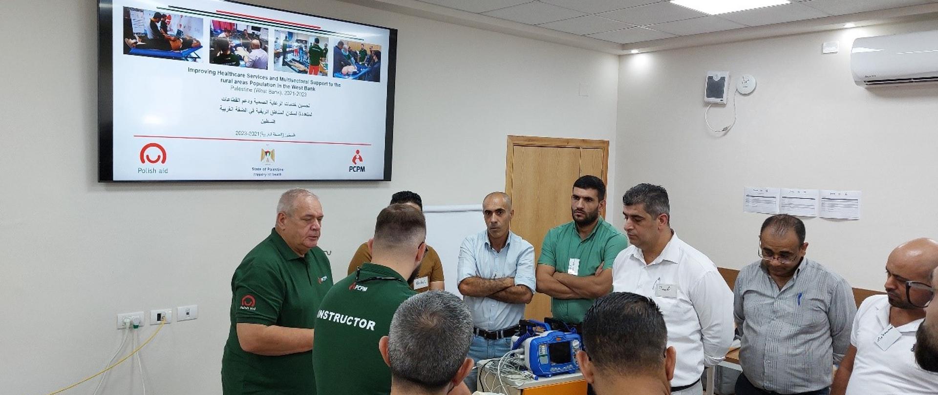 Grupa mężczyzn w pomieszczeniu stoi wokół szafki, na której stoi sprzęt elektroniczny. Na ścianie na monitorze widać zdjęcia logo Polskiej pomocy i PCPM. 