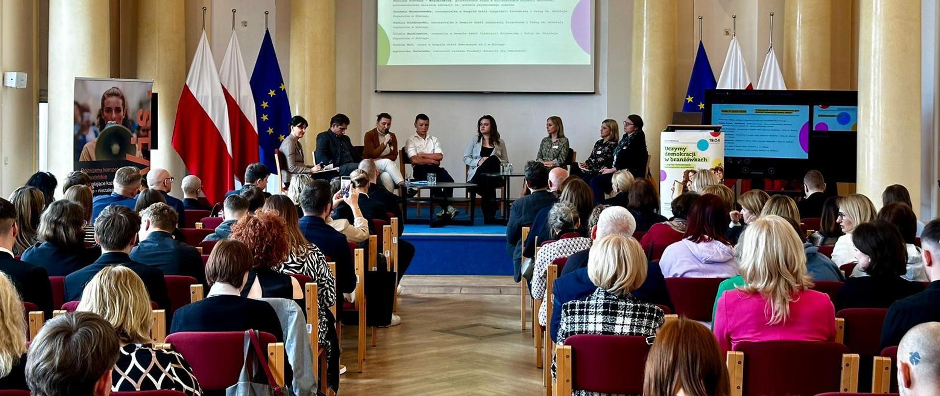 Na środku sali ustawiona jest scena, na której siedzi grono panelistów. Jednym z nich jest przemawiająca wiceminister Paulina Piechna-Więckieiwcz, która trzyma w ręku mikrofon. Za panelistami ustawione są flagi Polski i Unii Europejskiej, a nad nimi wisi ekran, na którym wyświetlane są podstawowe informacje o trwającym panelu. Przed sceną ustawionych jest kilka rzędów krzeseł, na których siedzą uczestnicy konferencji. 