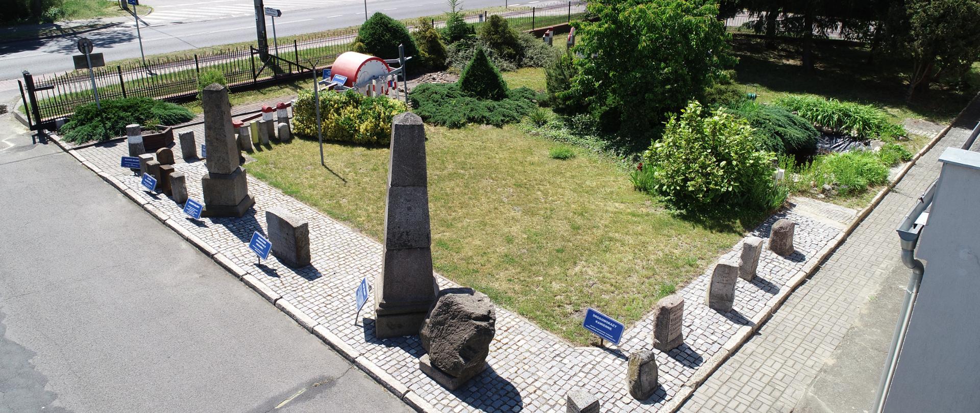 Zdjęcie z lotu ptaka. Na placu przed budynkiem ustawione eksponaty, w większości słupy milowe i graniczniki, a także walec konny. Przy eksponatach widnieją niebieskie tabliczki. Teren ekspozycji zagospodarowany zielenią. Z terenem placu graniczy droga krajowa.