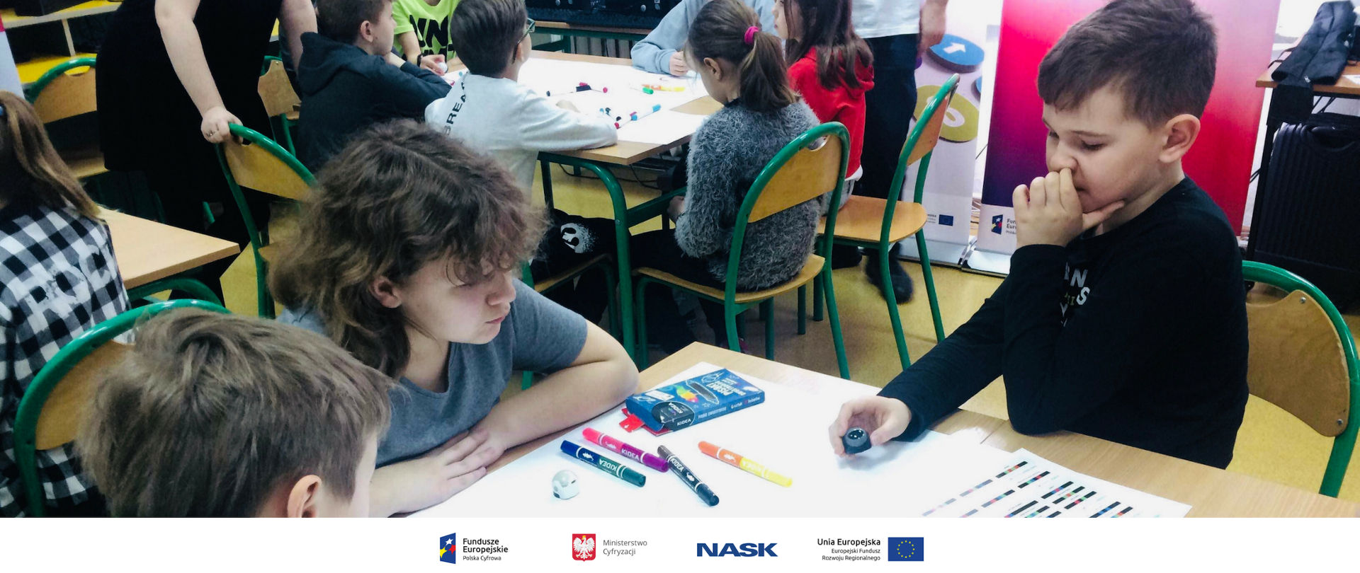 Na zdjęciu widać trzech chłopców przy szkolnej ławce. Na ławce rozłożone są kartki i flamastry. Na dole zdjęcia umieszczone są logotypy: Fundusze Europejskie. Polska Cyfrowa, Ministerstwo Cyfryzacji, NASK oraz Unia Europejska. Europejski Fundusz Rozwoju Regionalnego. 