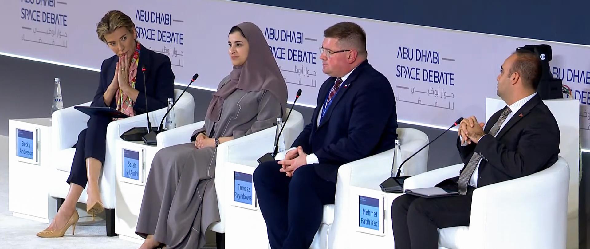 Dwie kobiety, jedna w szarym hidżabie, druga w granatowym ubraniu, i dwóch mężczyzn w garniturach siedzi na białych fotelach w rzędzie.