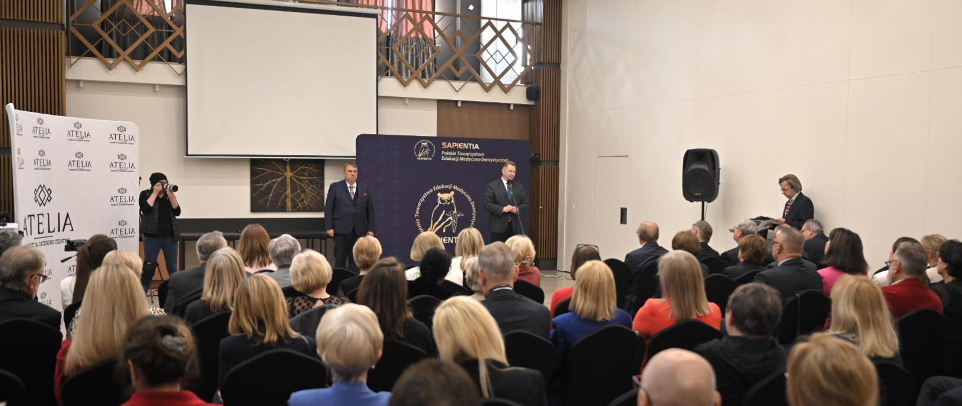 Widok z tyłu na wypełnioną ludźmi salę, z przodu stoi mężczyzna w garniturze i mówi do mikrofonu na stojaku, po obu jego stronach stoi dwóch mężczyzn, za nimi ciemnoszary baner, dalej wielki ekran, w tle widać biegnący dookoła ścian sali balkon.