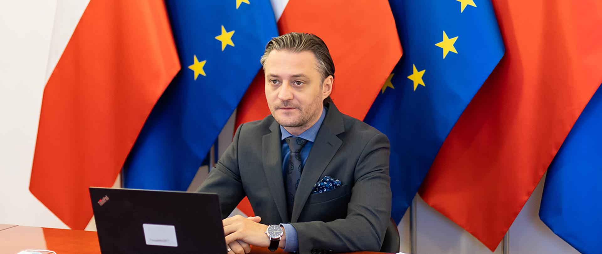 Wiceminister Bartosz Grodecki podczas wideokonferencji. W tle flagi Polski i UE