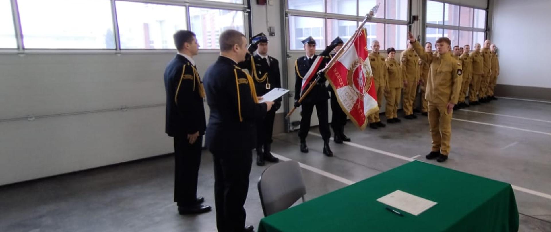 Zdjęcie przedstawia uroczysty moment składania ślubowania przez nowo przyjętego strażaka Pawła Przyborowskiego do służby w Komendzie Powiatowej Państwowej Straży Pożarnej w Gołdapi oraz sztandar komendy. W tle można zaobserwować pozostałych strażaków biorących udział w uroczystej zbiórce