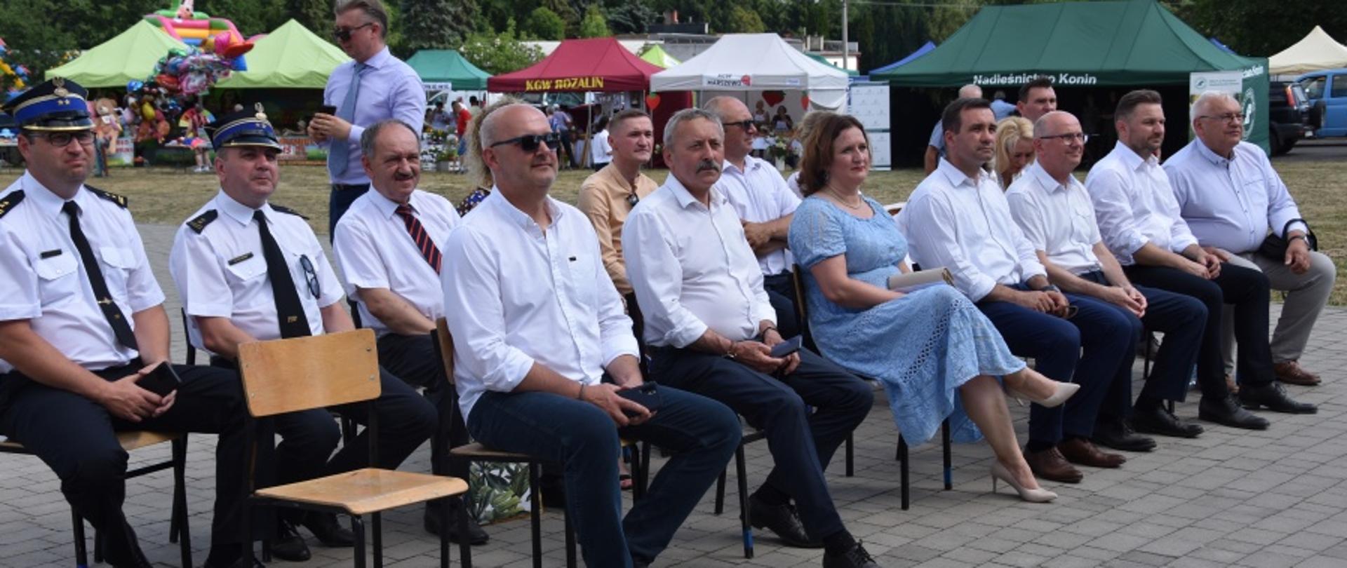 grupa ludzi, w tym strażacy w białych koszulach i rogatywkach siedzą na krzesłach, w tle kolorowe namioty