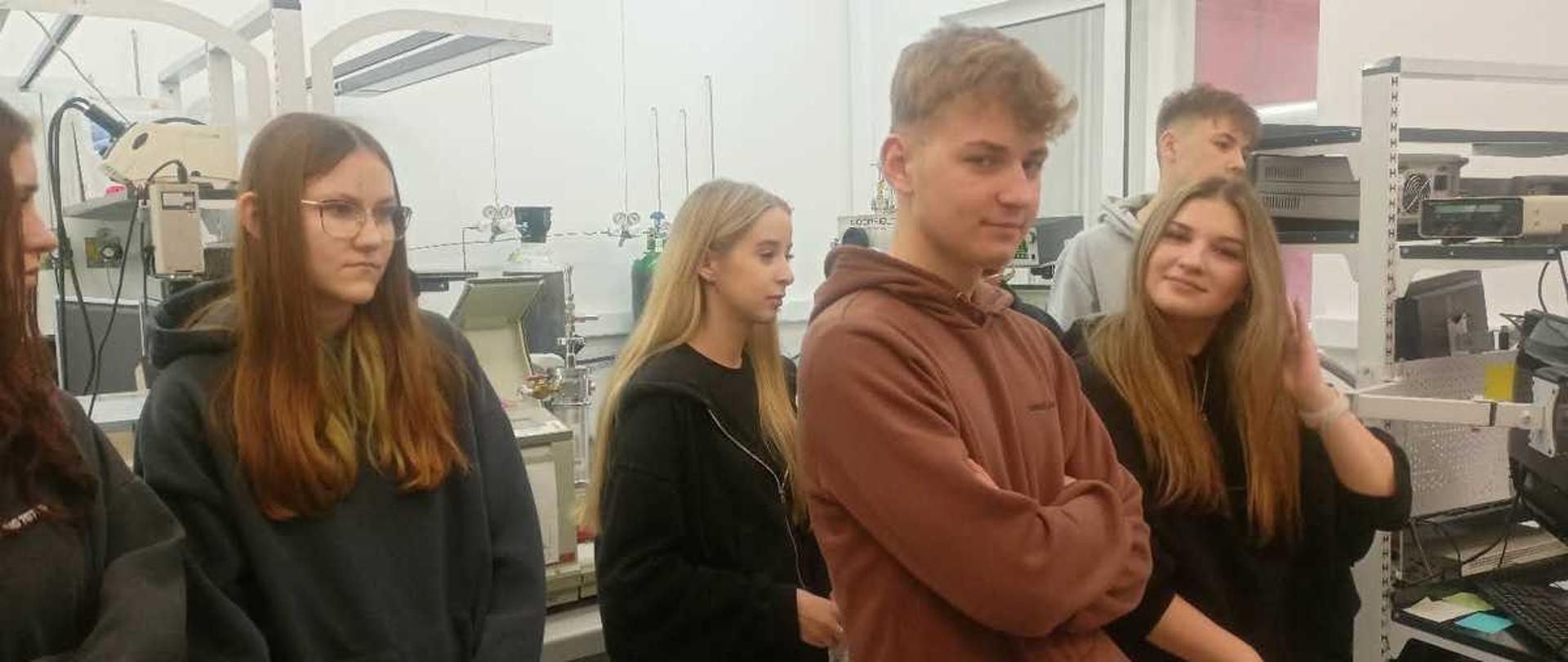 To zdjęcie przedstawia grupę młodych ludzi przebywających w pomieszczeniu przypominającym laboratorium naukowo-techniczne. W tym przypadku jest to Warszawska Akademia Techniczna.