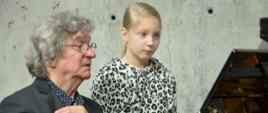 Profesor siedzi przy fortepianie, patrzy w stojące na pulpicie nuty, po prawej stoi dziewczynka we wzorzystej bluzce, patrzy w nuty