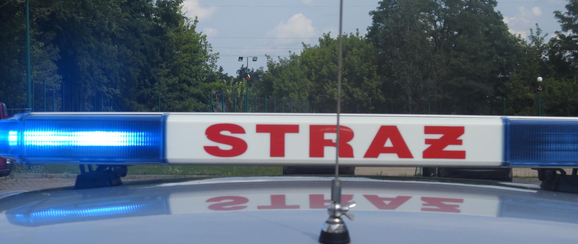 Dach samochodu z napisem straż i migającymi światłami. W tle widać niebo i korony drzew.