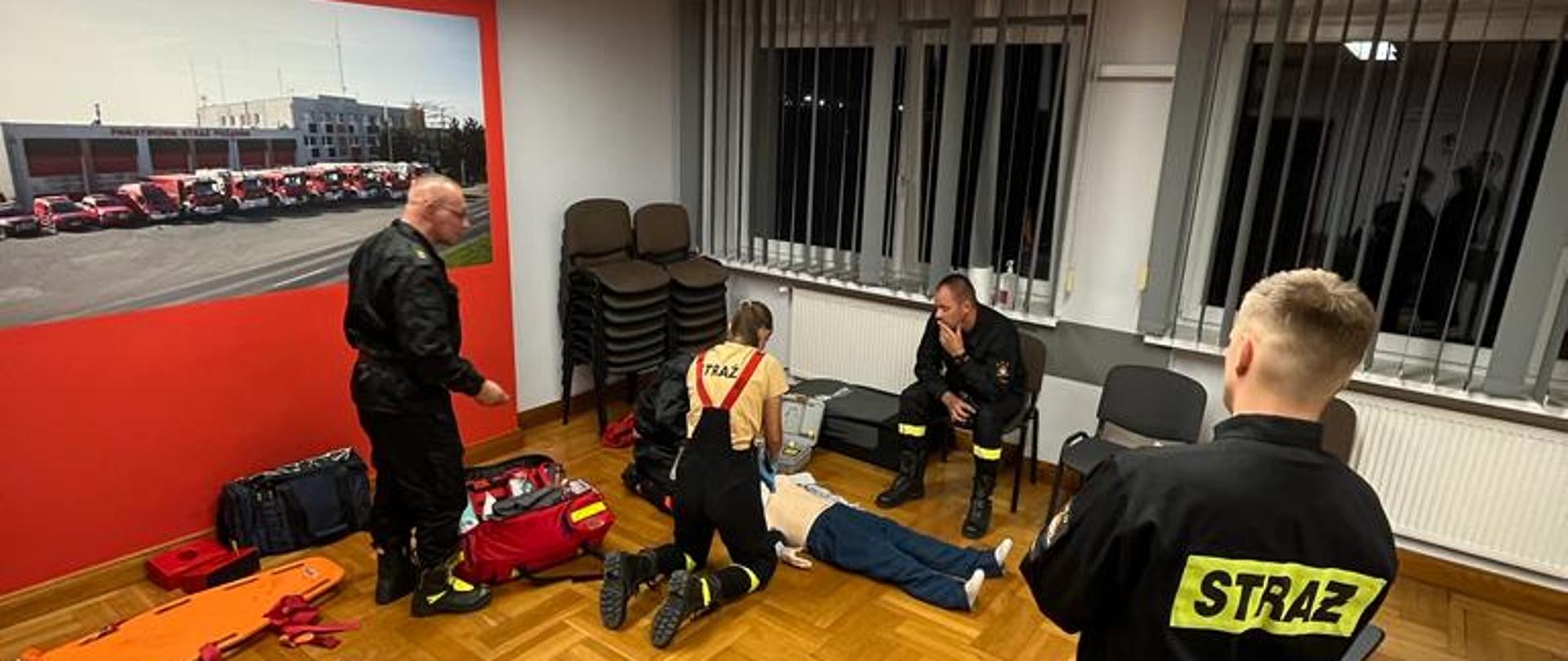 Strażacy w mundurach ćwiczą na manekinie w pomieszczeniu. Obok rozłożony sprzęt medyczny.