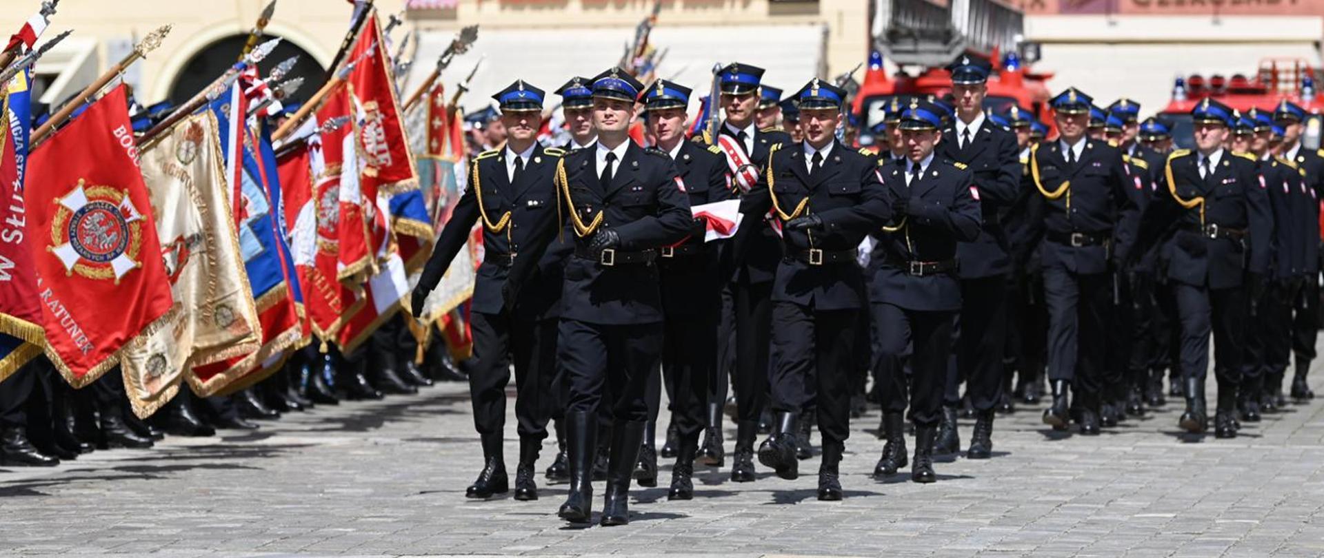 Kilkudziesięciu strażaków zgrupowanych w kolumnę marszową, w umundurowaniu reprezentacyjnym. W tle samochody strażackie i sztandary.