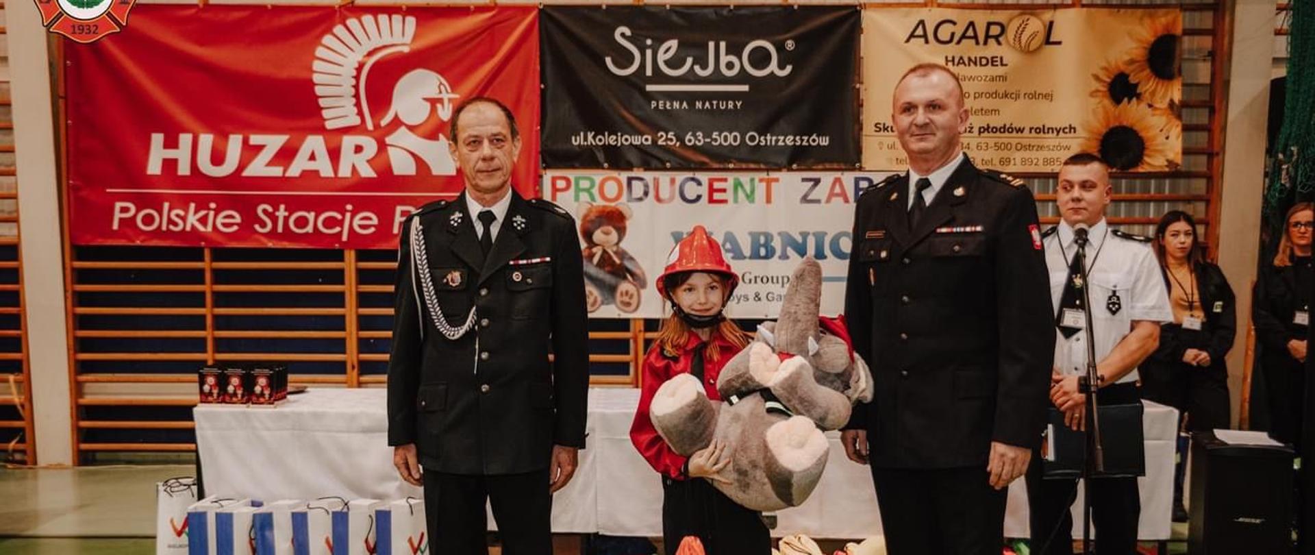 Prezes Zarządu Gminnego OSP oraz Komendant Powiatowy PSP wręczają nagrodę uczestnikowi zawodów. Zdjęcie wykonane w hali sportowej.